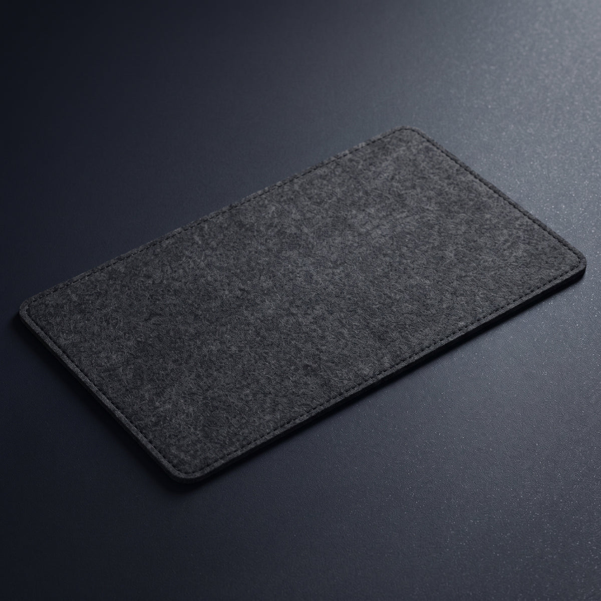 A dark gray felt desktop pad with rounded corners, shown at an angle on a dark gray textured background.