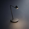 A modern black adjustable desk lamp with a circular base and curved arm, shown lit against a dark gray textured background.